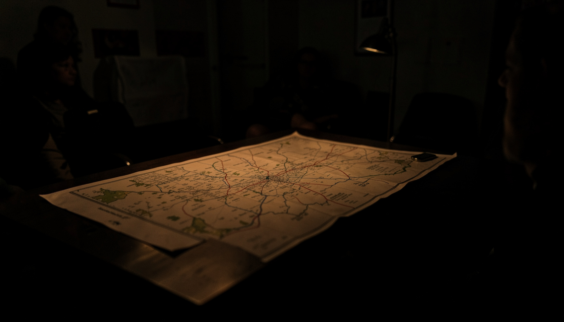 A dark, low-key photograph of a map spread across a table in a community meeting room, deep shadows with warm amber light pooling on the surface